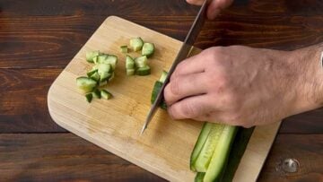 A hand chopping a cucumber with a knife on a wooden board.
