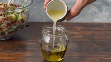 A hand pouring lemon juice in a jar with olive oil.