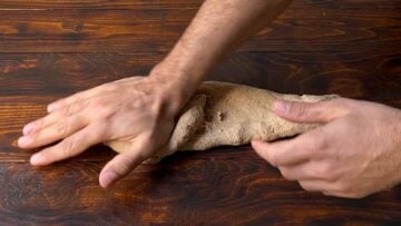 Two hands kneading barley bread dough on a wooden surface.