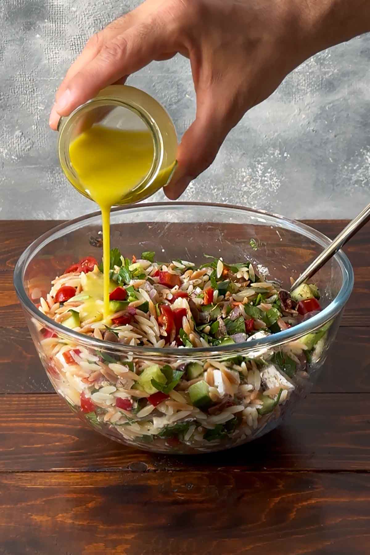 A hand pouring ladolemono dressing to a bowl with orzo salad.