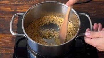 A hand stirring orzo with olive oil in a pot.