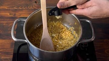 A hand adding minced garlic to the pot with orzo.