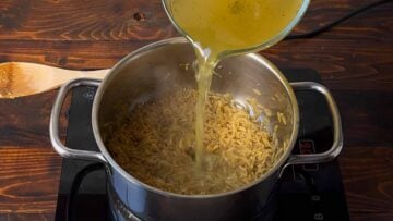 A hand pouring chicken broth to a pot with orzo.