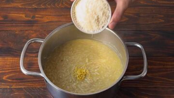 A hand adding Parmesan cheese to the pot with lemon orzo.