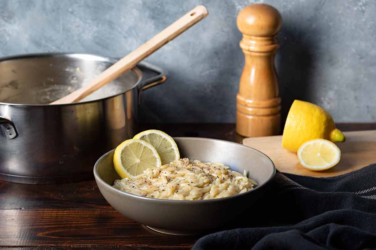 Lemon orzo in a bowl, a pot, a sliced lemon and a pepper grinder.