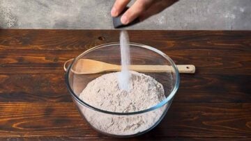A hand adding salt to a bowl with flour.