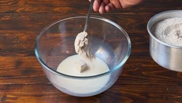 A hand adding flour to a bowl with milk and yeast.