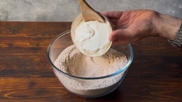 A hand adding Greek yogurt to a bowl with whole wheat flour.