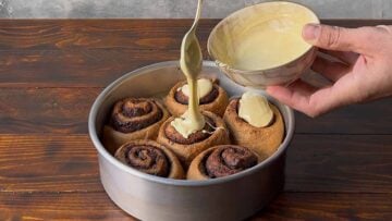 A hand spreading melted white chocolate on top of the cinnamon buns.