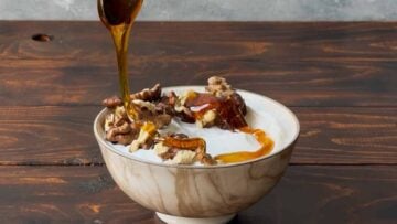 A hand adding honey to a bowl with Greek yogurt and walnuts.