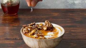 A hand sprinkling sea salt flakes to a bowl with Greek yogurt and honey.