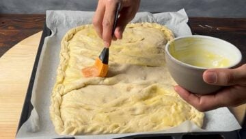 A hand brushing with butter the surface of the Greek cheese pie.
