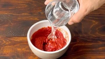 A hand pouring water to a bowl with tomato paste.