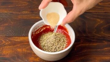 A hand adding onion powder to homemade pizza sauce.