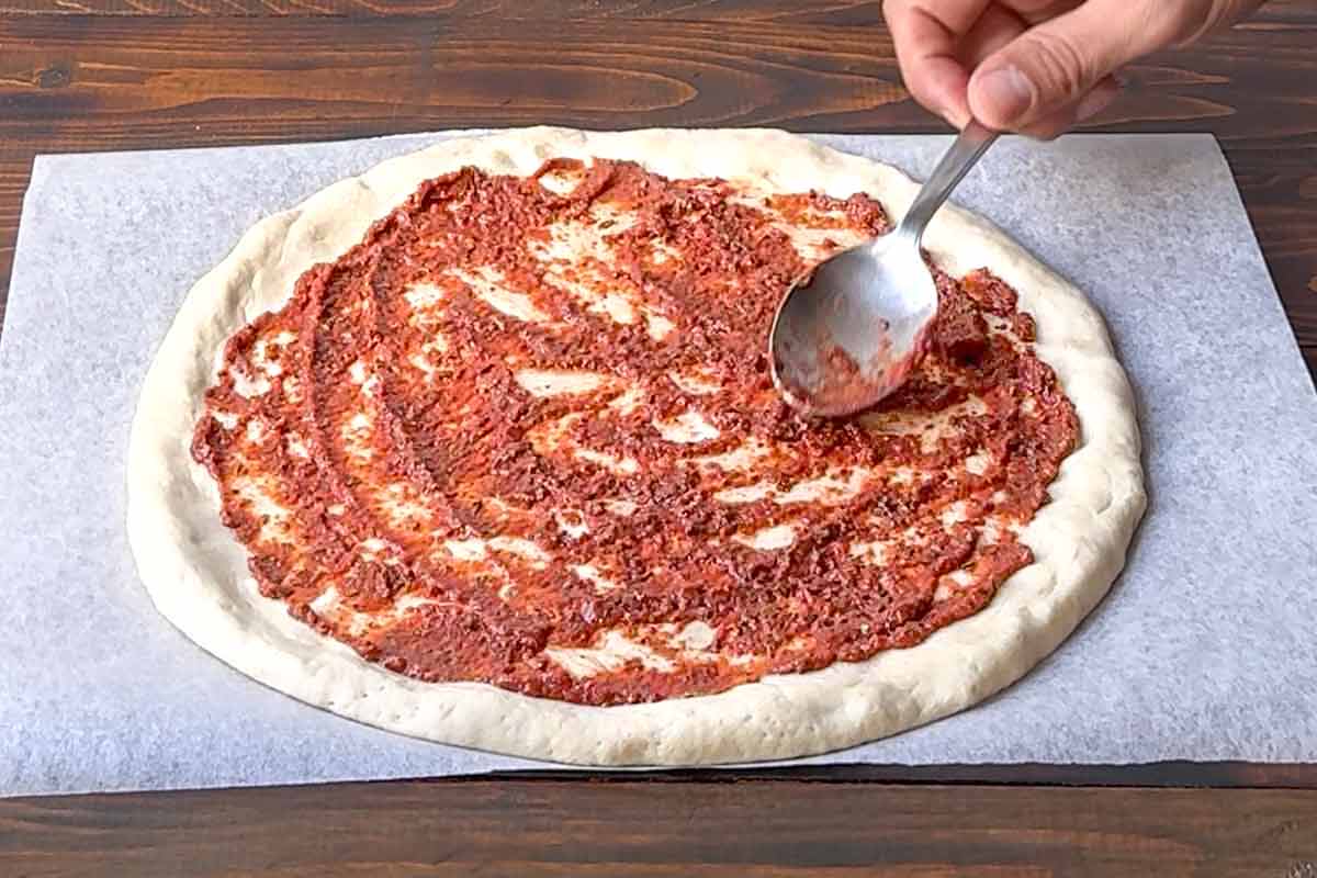 A spoon spreading tomato sauce over pizza dough.